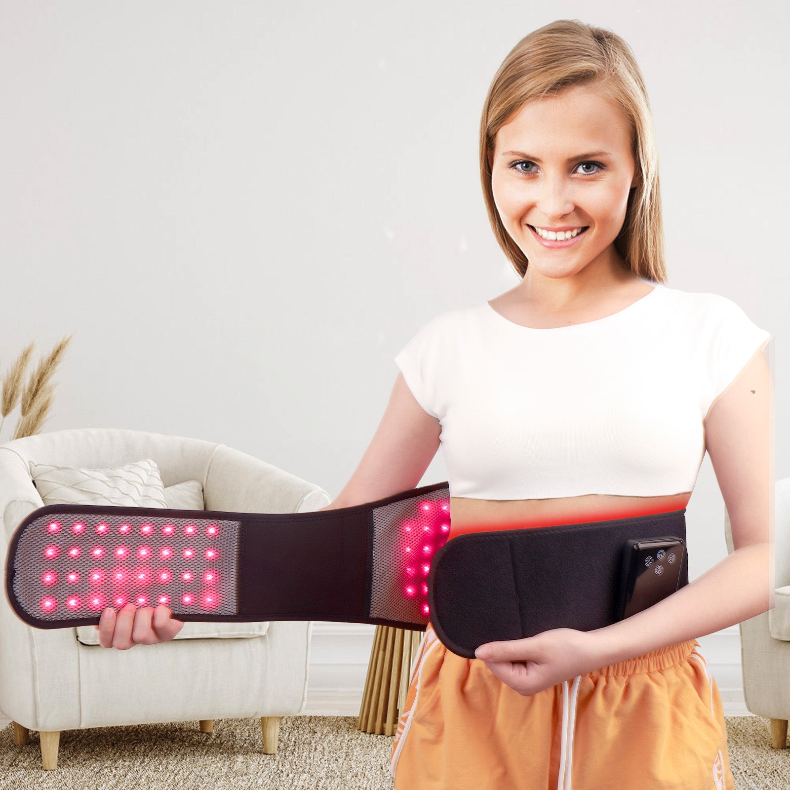 A woman wearing a white crop top and orange skirt holds up a black LED light therapy waist wrap device with glowing red lights, set against a bright, modern living room background.